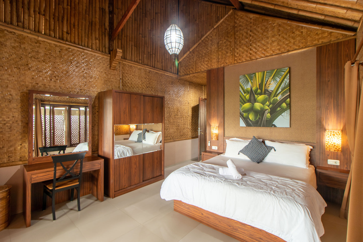 villa dengan kamar tidur yang bernuansa entik dengan dekorasi lampu gantung yang berada diatas tempat tidur dan lukisan pohon kelapa yang membuat suasana menjadi hangat serta lemari dan meja rias yang bisa kamu gunakan untuk menyimpan barang bawaan.