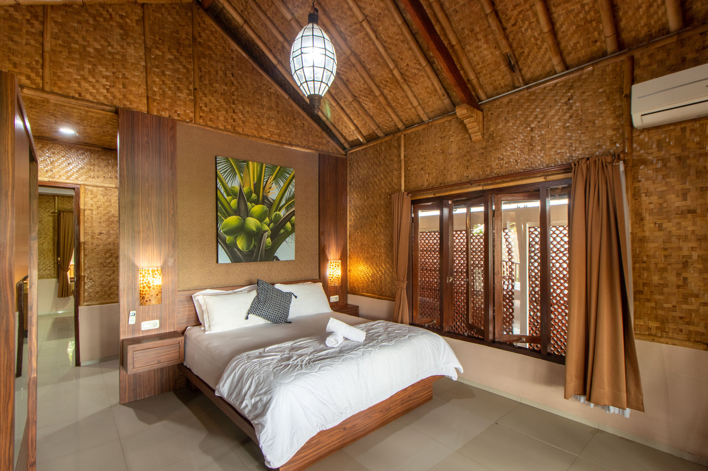 Kamar tidur Villa yang nyaman dengan kasur yang bisa muat untuk 2 orang, bukaan kaca yang cukup lebar, dihiasi lukisan bergambar pohon kelapa dan lampu gantung serta dilengkapi kamar mandi dalam dengan water heater.