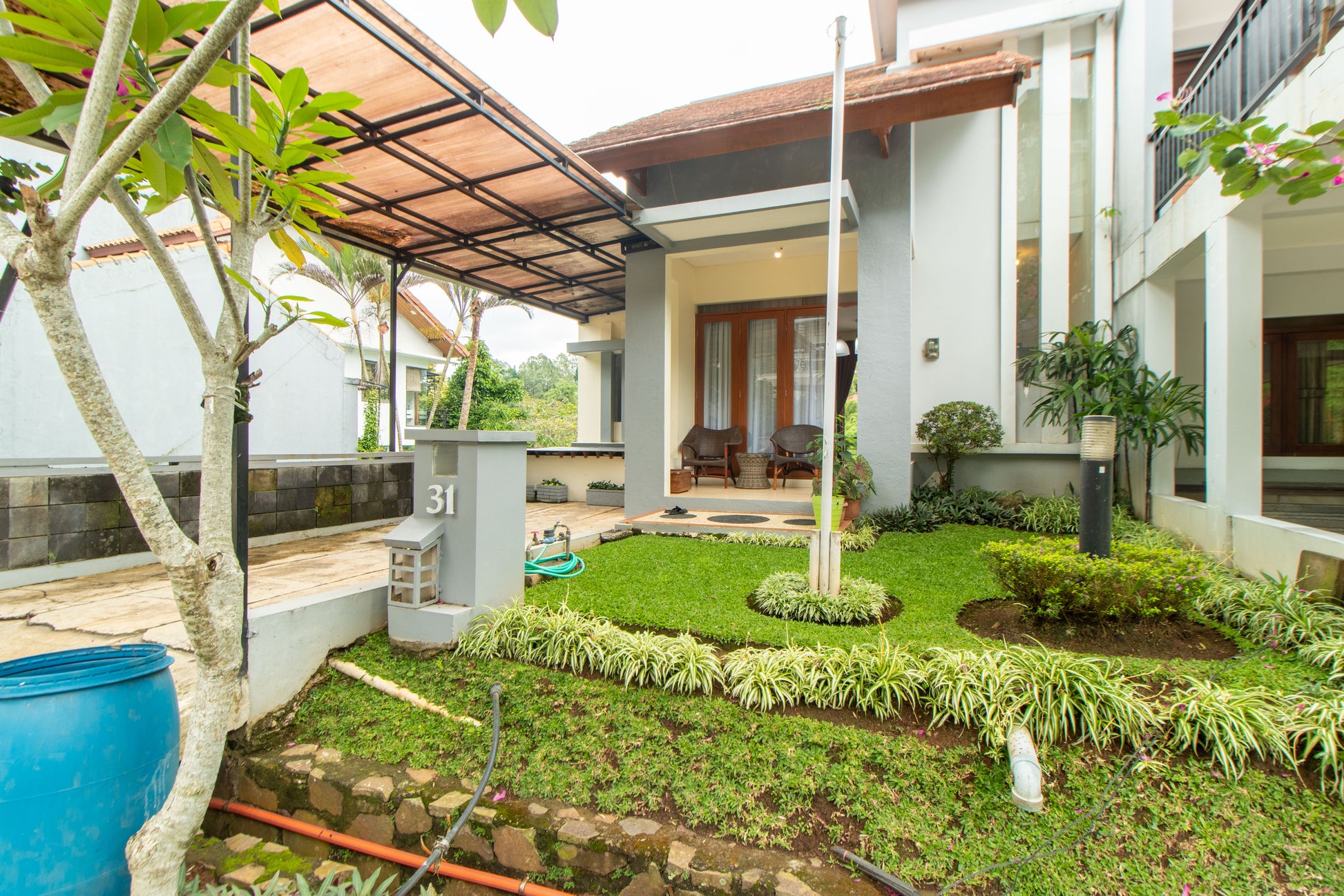 Taman depan yang ditumbuhi rumput hijau dan pohon menambah kesejukan di udara sekitar Villa, carport yang sudah ditutup kanopi dan tersedia 2 kursi santai dan meja di depan Villa.