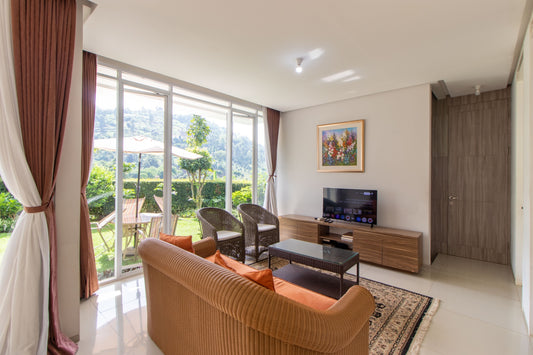 Ruang tamu yang luas dan terang di sebuah vila yang berlokasi di dago Bandung dengan sofa besar berwarna oranye, pintu kaca yang menuju ke balkon, dan TV layar datar di atas dudukan lemari kayu.