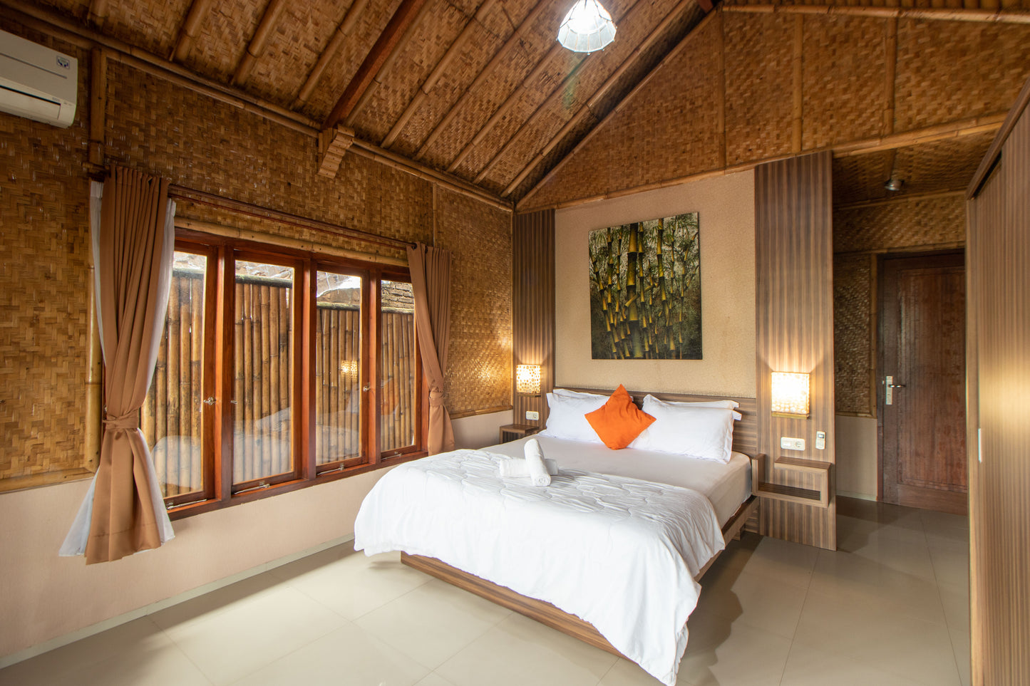 Villa dengan Kamar tidur yang konsep bambu dilengkapi dengan kasur berukuran besar dengan lampu tidur diatas kepala ranjang, lukisan bambu dan lampu gantung unik