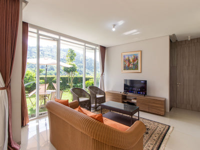 Ruang tamu yang luas dan terang di sebuah vila yang berlokasi di dago Bandung dengan sofa besar berwarna oranye, pintu kaca yang menuju ke balkon, dan TV layar datar di atas dudukan lemari kayu.